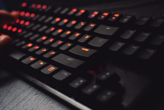 Tastatură de gaming cu taste luminate, lângă un monitor pe care rulează un joc