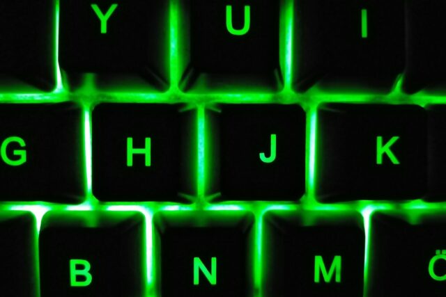 Monitor luminat într-o cameră întunecată, cu o tastatură și o cană pe birou seara târziu